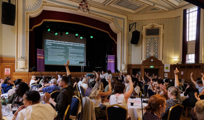 People sat at tables in large hall raising hands and looking at a person in front of the stage