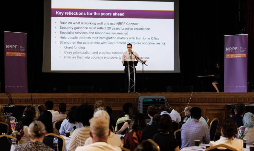 Person standing on stage in front of slide speaking to room of people
